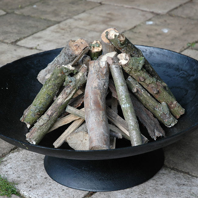 A black metal fire pit filled with neatly stacked logs, ready to be lit. The scene is outdoors, set on a stone-paved patio.